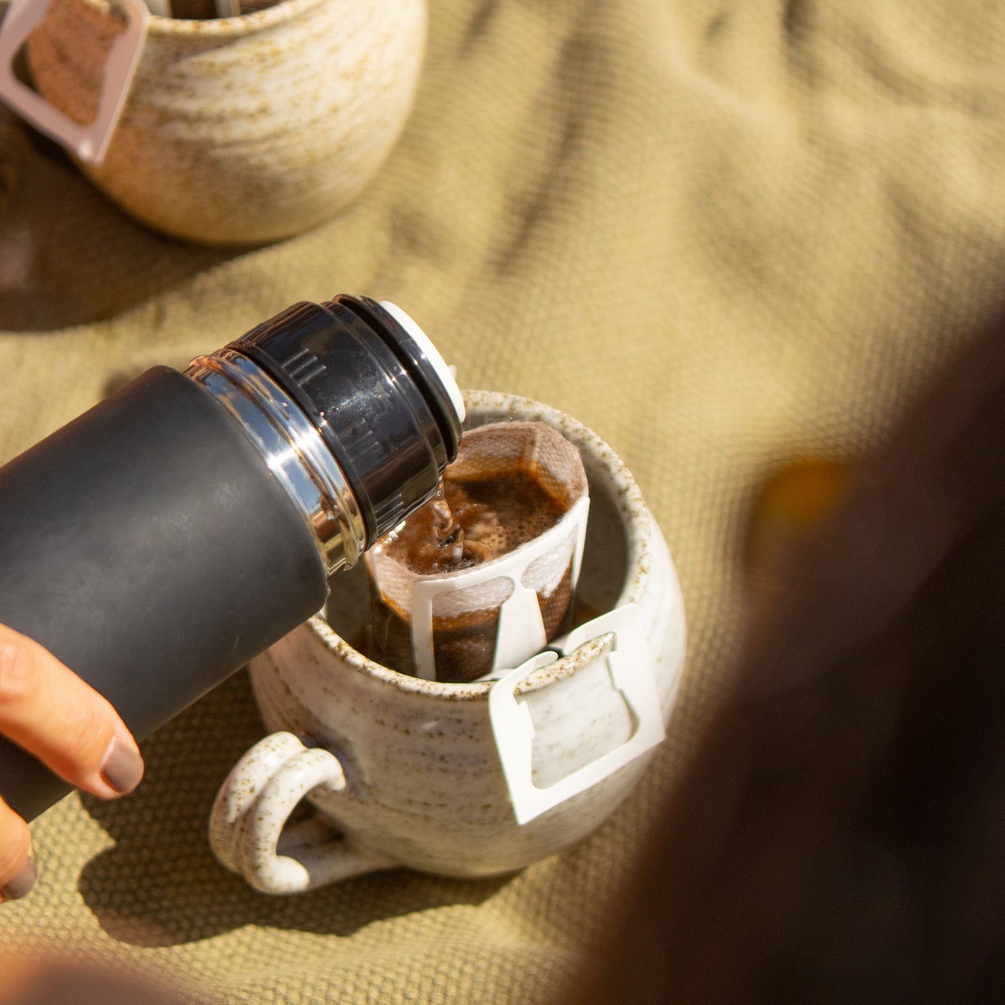 Hand pours hot water into Field Drip single-serve drip coffee bag over a ceramic mug on an outdoor picnic blanket