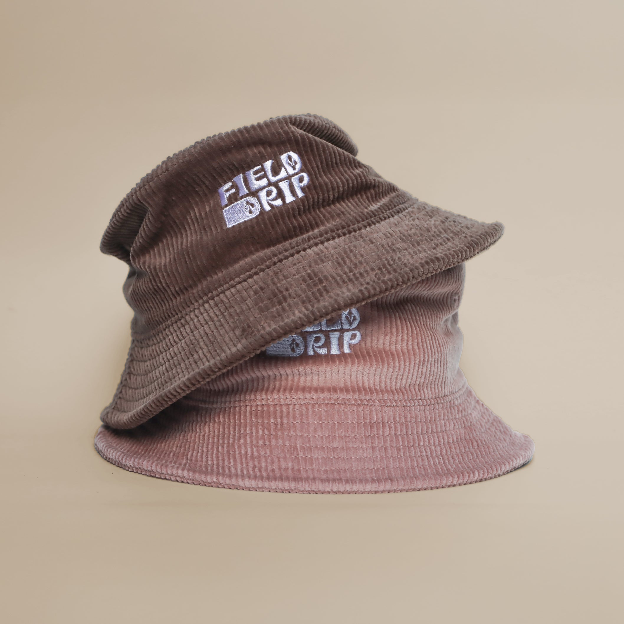 Two Field Drip corduroy bucket hats—brown and hazy pink—stacked, showing the embroidered logo on the front.