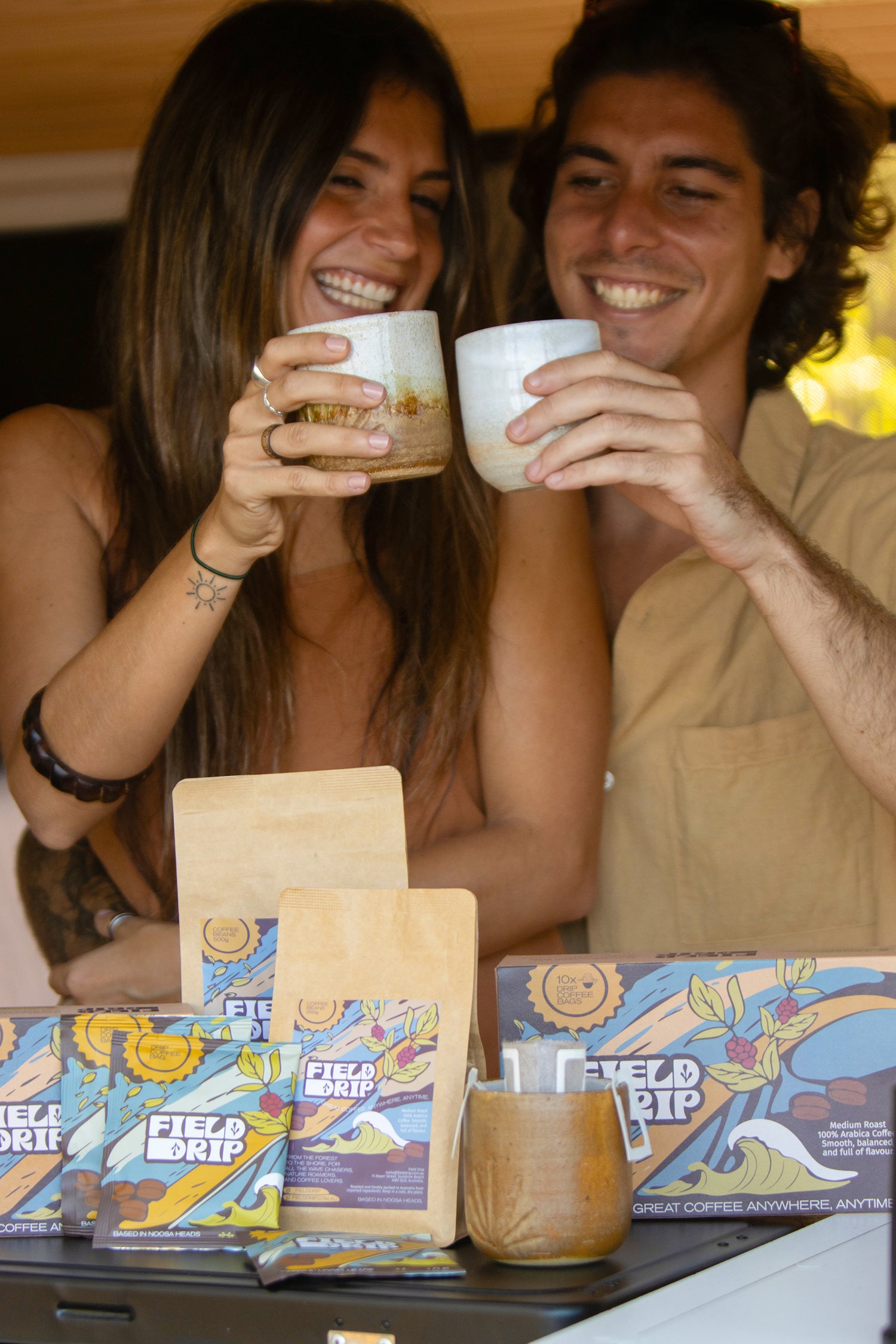 Couple cheers ceramic cups beside Field Drip specialty coffee bags and drip box in a van, Sunshine Coast Australia.