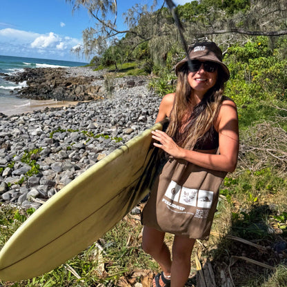 Woman at a rocky beach carrying a surfboard, wearing a Field Drip bucket hat and walnut cotton tote bag with printed logo.