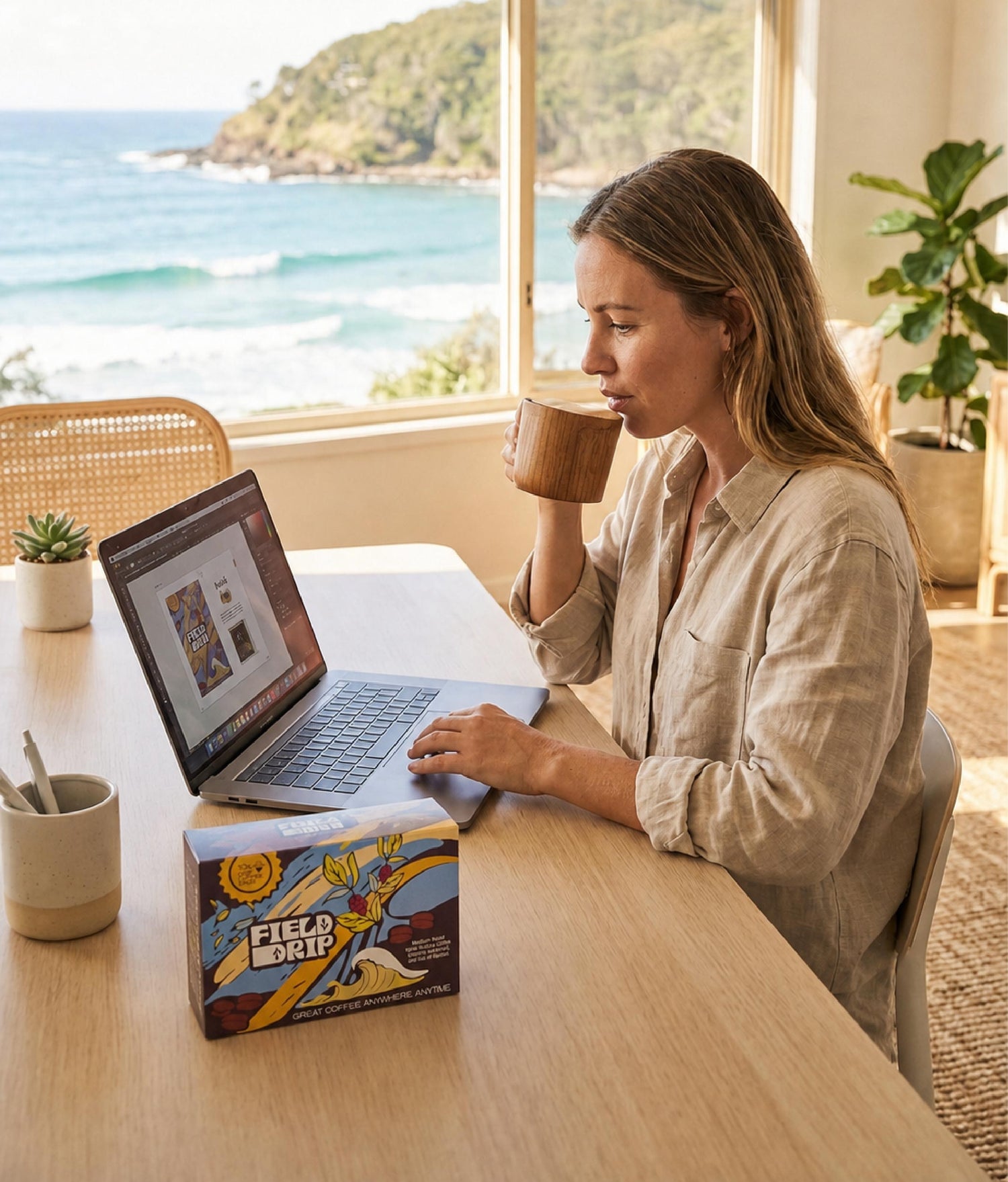 Woman drinking from a mug and using a laptop at a desk with a scenic view outside
